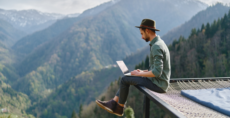 ein Mann mit Hut sitzt in den Bergen und arbeitet auf einem Laptop