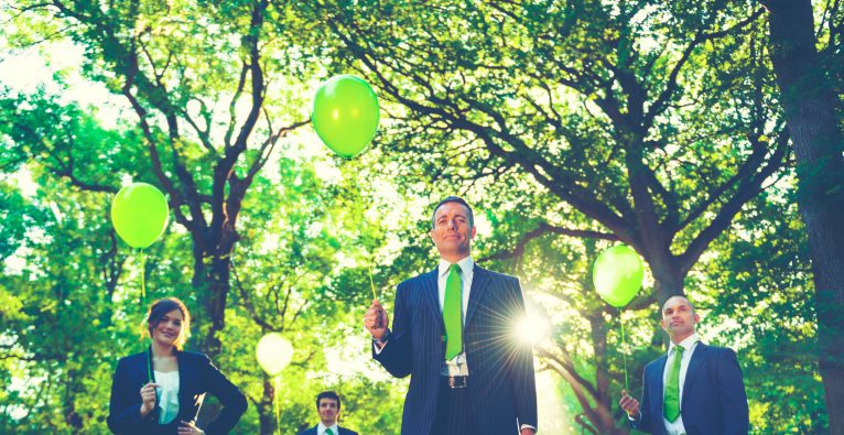 Personen in Anzügen stehen in einem Wald und halten grüne Luftballons.
