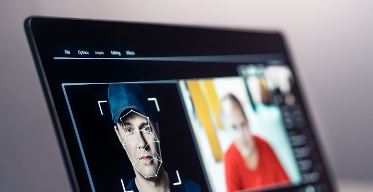 Computerbildschirm zeigt auf der linken Seite des Bildschirms das Gesicht eines Mannes mit einer Mütze zu sehen, auf das ein geometrisches Netz projiziert ist. Rechts im Bild ist ein unscharfer Ausschnitt einer anderen Person zu erkennen.