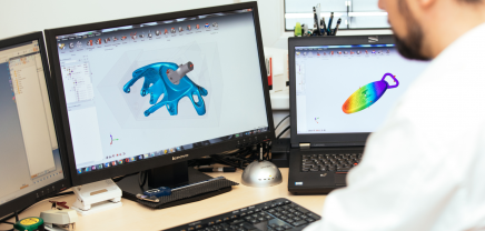 Das Bild zeigt eine Person, die an einem Schreibtisch vor zwei Computerbildschirmen arbeitet. Auf dem linken Bildschirm ist ein 3D-Modell eines blauen mechanischen Teils zu sehen. Auf dem rechten Bildschirm wird ein weiteres 3D-Modell mit einer bunten Analyseanzeige dargestellt. Die Person trägt ein weißes Hemd und ist nur teilweise von hinten zu sehen. Auf dem Schreibtisch befinden sich verschiedene Büromaterialien wie ein Hefter, ein Maßband und eine externe Festplatte. Die Szene vermittelt eine Arbeitsumgebung im Bereich CAD (Computer-Aided Design) oder technisches Design.