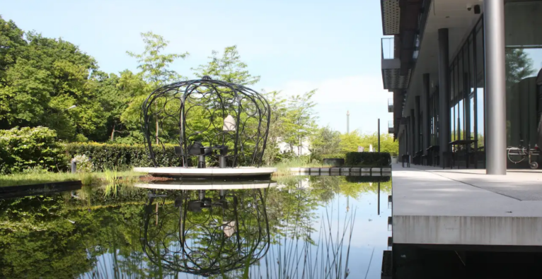 Veranstaltungsort mit einer Terrasse, die an einen großen Teich grenzt. Im Vordergrund ist eine Sitzgelegenheit zu sehen, die auf einer runden Plattform im Wasser liegt. Die Umgebung ist von Grün und Bäumen umgeben