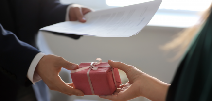 Eine Person in einem Anzug hält ein Dokument in der Hand und überreicht einer anderen Person ein kleines, quadratisches Geschenk, das in rosa Papier eingewickelt und mit einem weißen Band verziert ist. Die Szene findet in einem Büro statt und beide Personen sind nur teilweise sichtbar.
