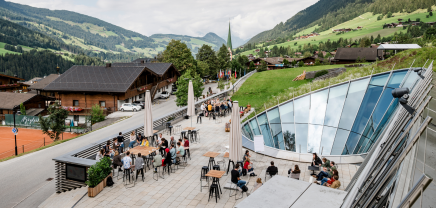 Schauplatz des Europäischen Forums Alpbach - mit Blick auf den Ort Alpbach. Ein asphaltierter Platz, Menschen versammelt umgeben von modernem Glas und grünen Almwiesen.