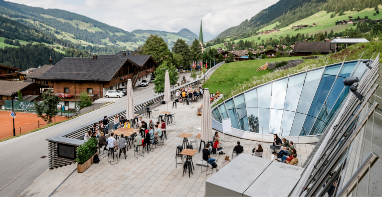 Schauplatz des Europäischen Forums Alpbach - mit Blick auf den Ort Alpbach. Ein asphaltierter Platz, Menschen versammelt umgeben von modernem Glas und grünen Almwiesen.