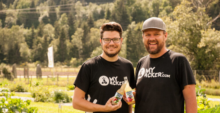 Die myAcker CEOs Patrick Kleinfercher und Christoph Raunig stehen in einem Garten und halten ihren Ackersaft in den Händen.