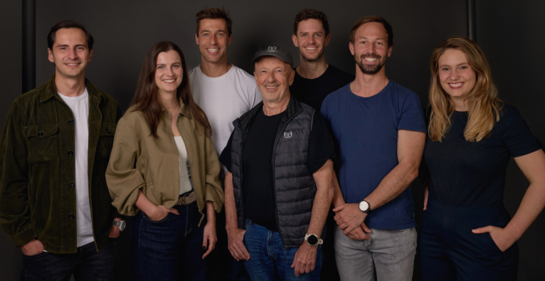 Hansi Hansmann und die Gründer von one100 stehen vor einer dunklen Wand und lachen in die Kamera.